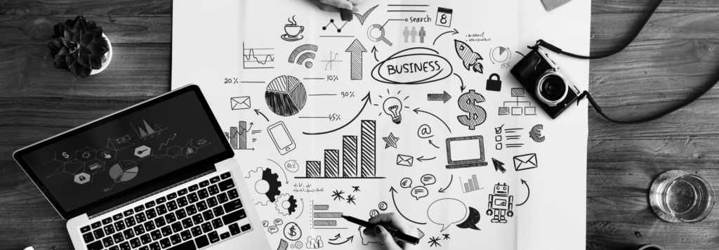 A top-view workspace featuring a laptop, a camera, and a brainstorming sheet with hand-drawn business strategy icons, including charts, graphs, and digital marketing elements.